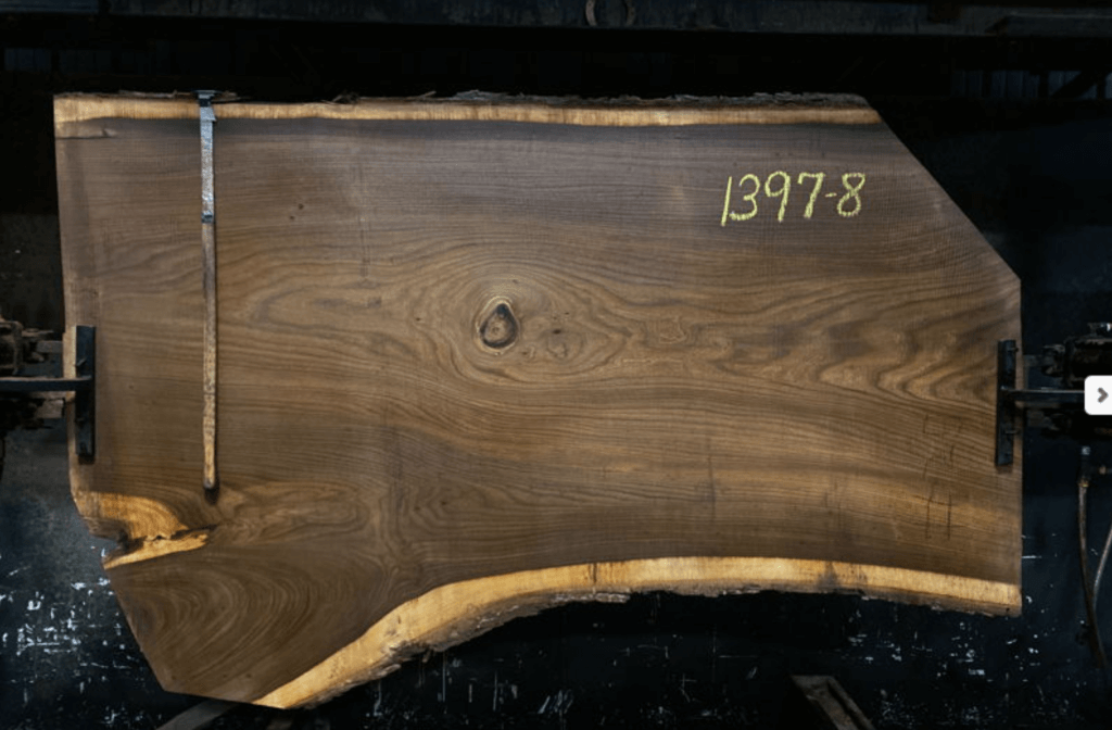 A large, rustic elm slab with a rough, natural edge is marked with the number "1397-8" in yellow chalk. The plank is vertically positioned and secured by clamps on either side. The rich wood grain of this Slab #1397-8 is clearly visible against a dark background.
